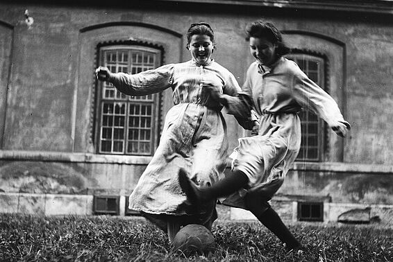 Zwei junge Frauen in altmodischen Kleidern spielen lachend Fußball. Schwarzweiß-Fotografie