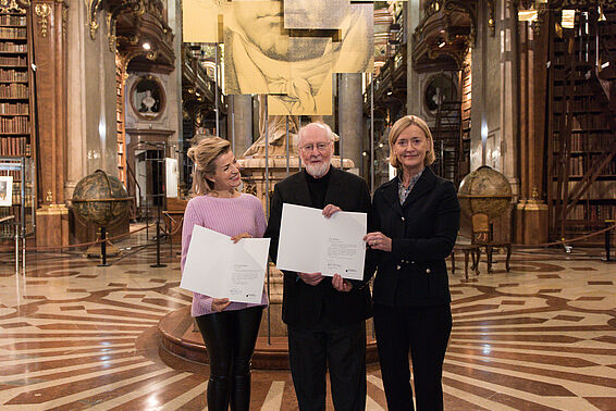Zwei Frauen und ein Mann halten zwei Urkunden in einem Marmorsaal