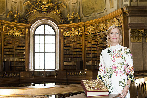 Foto von Frau die lächelnd mit einem prachtvollen, alten Buch in einem Saal voller Bücherregalen posiert
