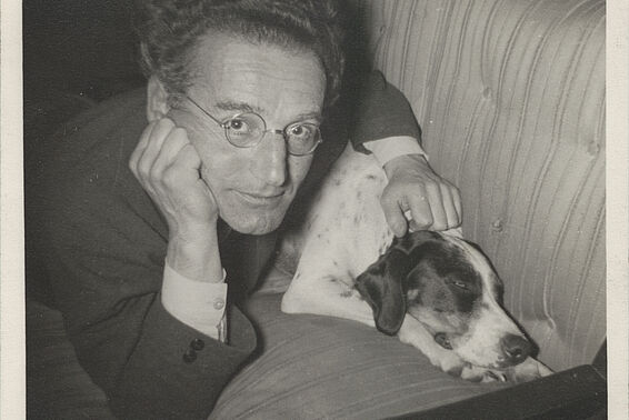 Schwarz-weißes Foto von Mann mit Brille, der einen Hund auf einer Couch streichelt und in die Kamera blickt