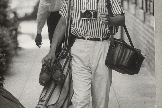 Ganzkörper-Portrait eines lächelnden Mannes in gestreiftem Hemd mit mehreren altmodischen Kamerataschen, schwarz-weiß
