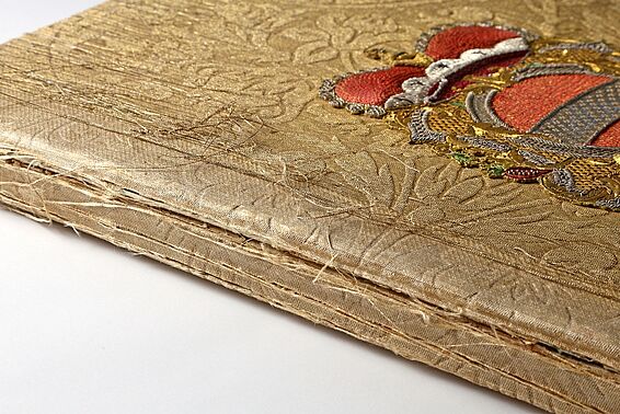 Foto von mit goldenem Stoff eingebundenem, alten Buch mit Wappen