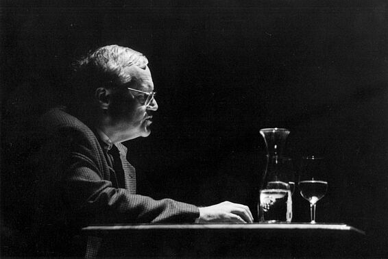 Mann mit Brille sitzt an einem Tisch und spricht, nur von oben beleuchtet, schwarz-weiß