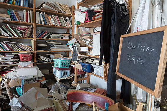 Unordentliches Zimmer mit vollen Bücherregalen, rechts hängt eine Schiefertafel mit den Worten "hier ALLES TABU"