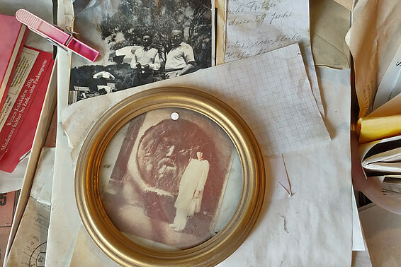 Zettel- und Fotostapel auf einem Tisch liegend. Das oberste Bild ist golden eingerahmt und zeigt ein altes Foto einer Frau, die neben einer Statue steht