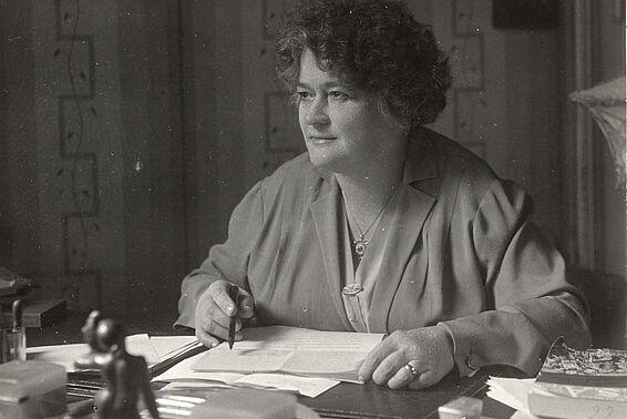 Frau mit gelockten Haaren sitzt an einem Tisch und schreibt, den Blick in die Ferne gerichtet, schwarz-weiß