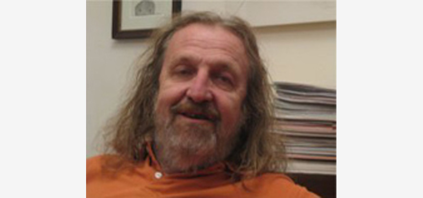 Mann mit langen Haaren und Bart, orangem Oberteil vor Papierstapel
